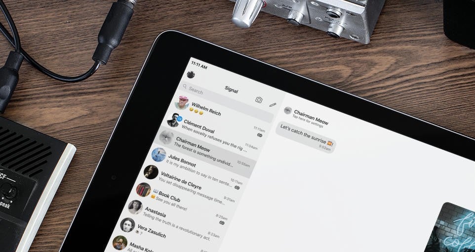Un iPad en la parte inferior derecha que muestra la versión iOS de la aplicación Signal con una lista de personas a la izquierda y una pantalla de chat a la derecha, con otros dispositivos y cables parcialmente visibles.
