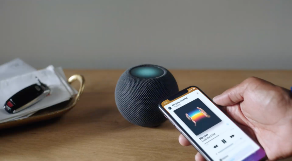 Un mini Homepod noir debout sur une table en bois et un smartphone jouant de la musique tenue en main à côté